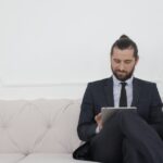 A businessman in a suit sitting on a sofa using a tablet, smiling.